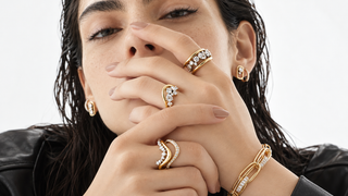 Close up portrait of a woman wearing sculptural yellow gold and diamond rings, matching earrings and a statement bracelet, with her hands posed elegantly near her face.