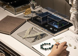 A designer working on a luxury jewelry sketch surrounded by gemstones, tools, and finished high-end pieces.