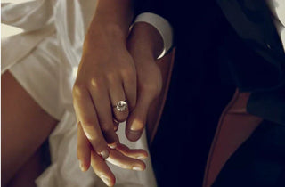 Close-up of two hands intertwined, one wearing a large round solitaire diamond engagement ring on a silver band, with a white dress and dark suit visible in the background.