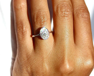 Close-up of a hand wearing a bezel set oval moissanite ring in white gold, reflecting rainbow light in direct sunlight