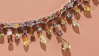 An elaborate high jewellery necklace featuring fancy coloured diamonds in shades of yellow, orange, champagne and brown, with pear, marquise and square cut stones suspended against a soft peach background.